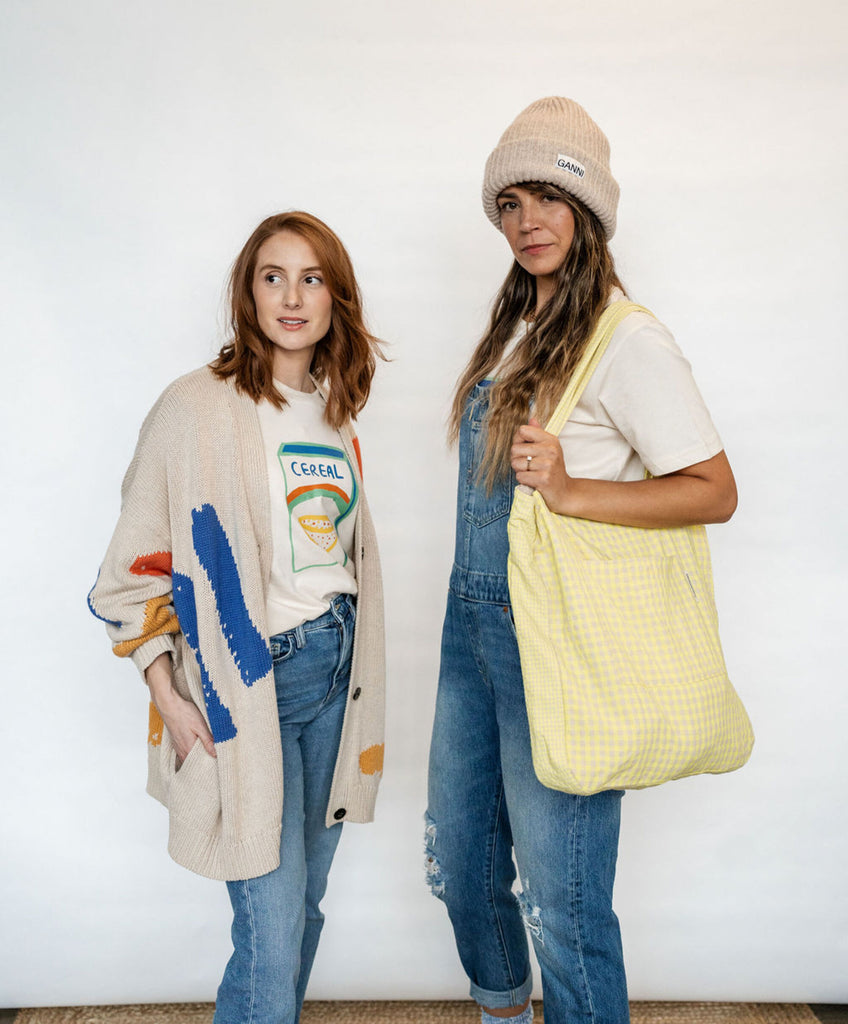 Two white woman stand next to each other. The woman on the left looks away from the camera and wears a t-shirt with Cereal drawing on it and a knitted cardigan with colourful motifs, from Cub & Pudding. The woman on the right wears a beanie hat from Ganni and holds a gingham yellow tote bag over her shoulder.