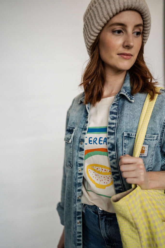 A white woman stands with a beanie hat on, a denim jacket and a t-shirt with a Cereal illustration on it from Cub & Pudding. She holds a yellow gingham bag over her shoulder.