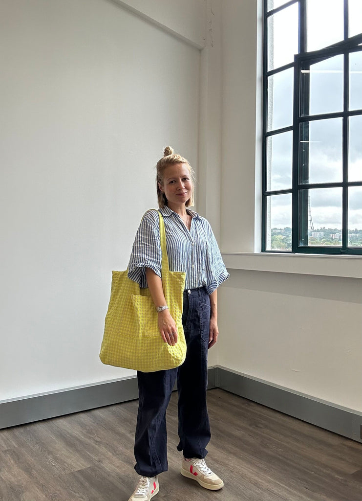 A white woman with topknot bun stands by a window smiling with a yellow gingham tote bag over shoulder from Cub & Pudding